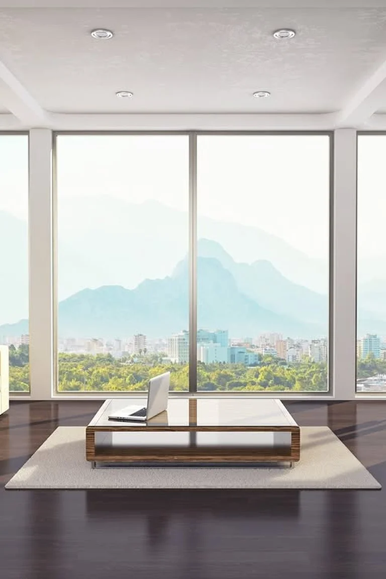Bright living room with floor-to-ceiling windows, cream sofa and laptop on the coffee table in soft sunlight.