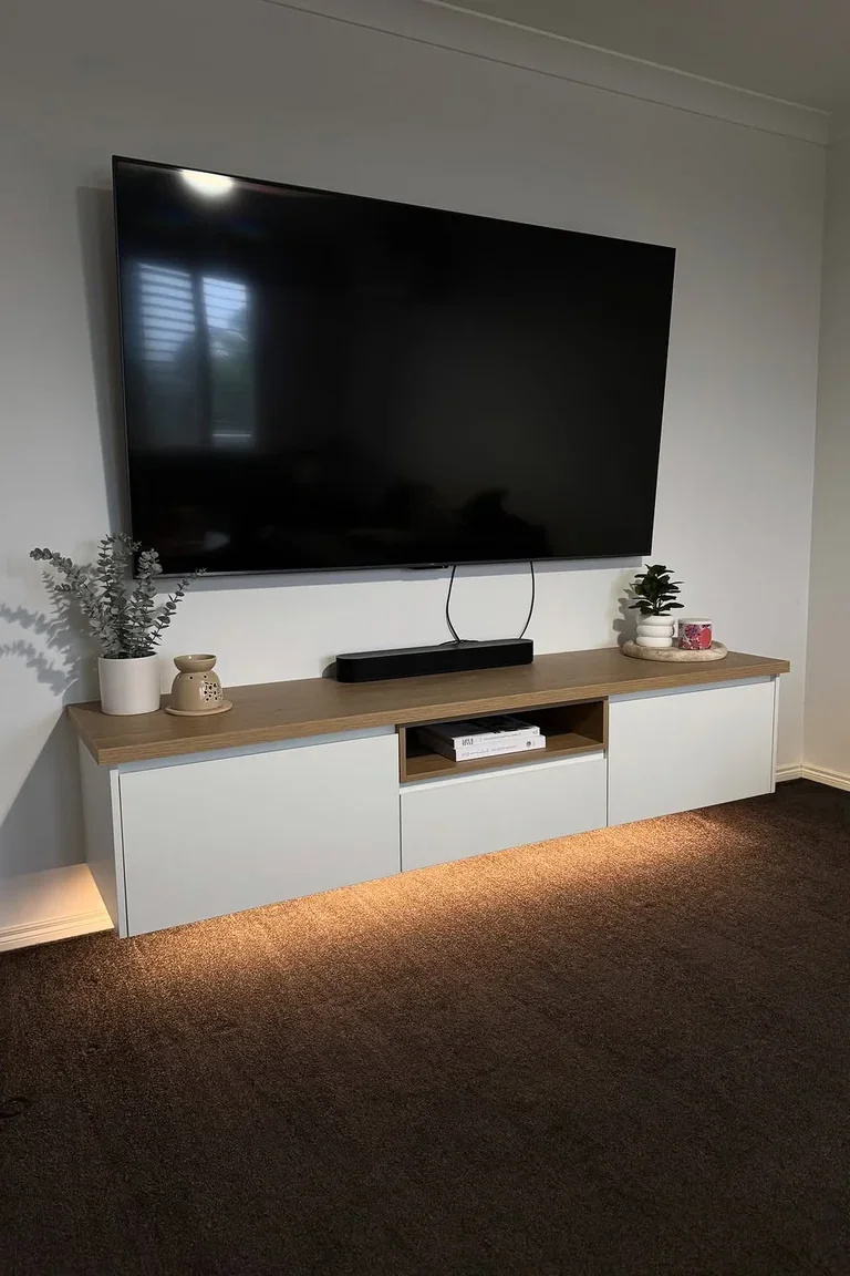 A minimalist living room TV wall with a plain white wall, a large wall mounted television above a long floating console with white handleless drawers and a wood top, a soundbar and small plants.