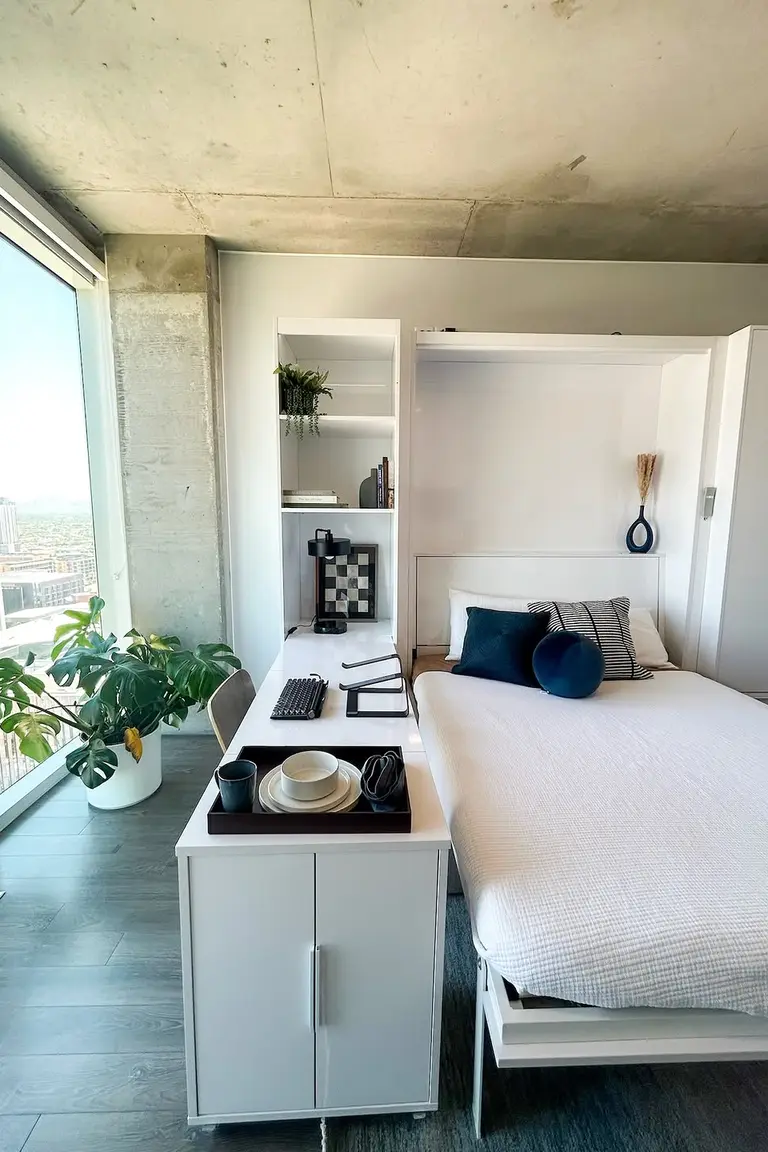 11 Micro Studio Apartment Ideas for Extremely Small Urban Spaces 3 White Murphy bed lowered beside a desk unit with shelves, books, and a plant, by a large window