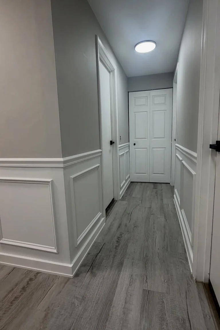 Hallway with white picture-frame wainscoting, soft gray upper walls, and wood-look flooring
