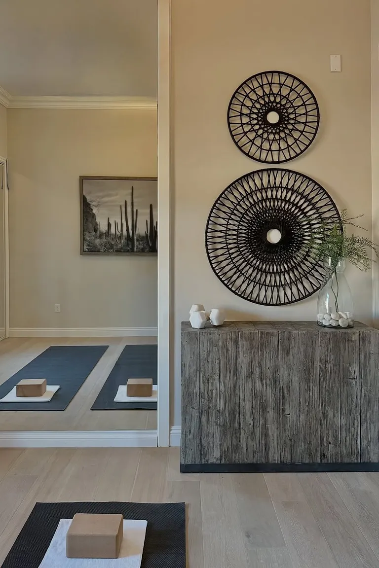 A calm yoga room with black mats and cork blocks on light wood flooring, a large wall mirror, a weathered wood cabinet, and round woven wall decor
