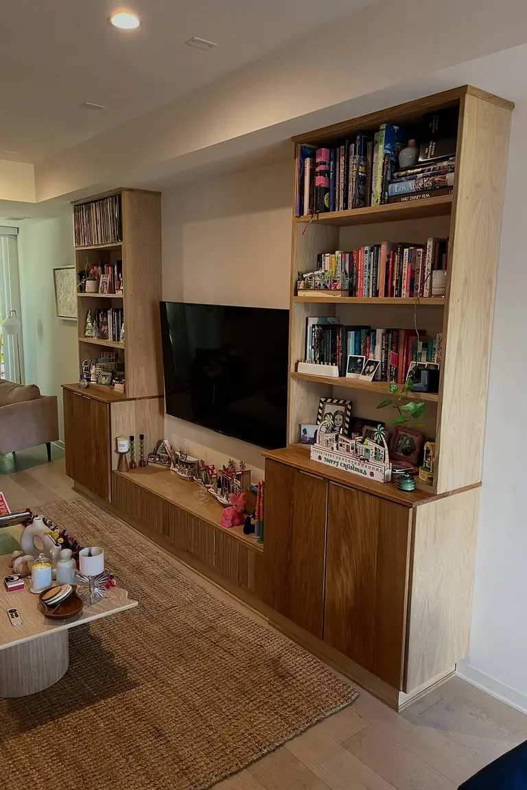 A living room TV wall with light oak built-in bookcases filled with books on both sides of a wall mounted television, a long low bench unit below, a rug, a coffee table and an armchair.