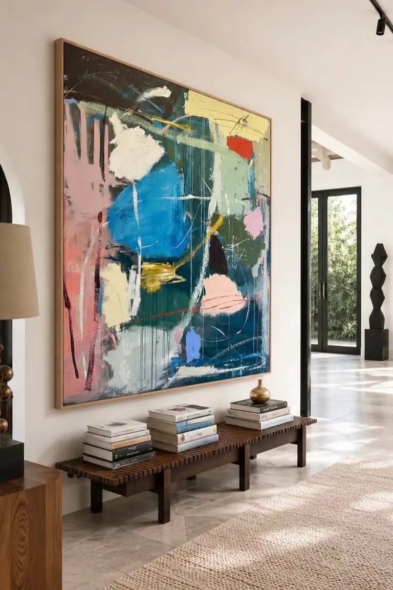 A bright hallway with a very large colorful abstract painting above a low dark wood slatted bench holding stacks of books, with a rug, arched doorway, and built-in shelves visible