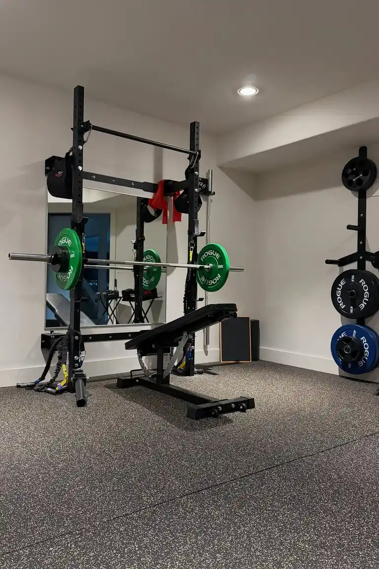 A home strength area with a black wall-mounted power rack holding a loaded barbell with green plates, an adjustable bench, a large wall mirror, plate storage, and speckled rubber flooring