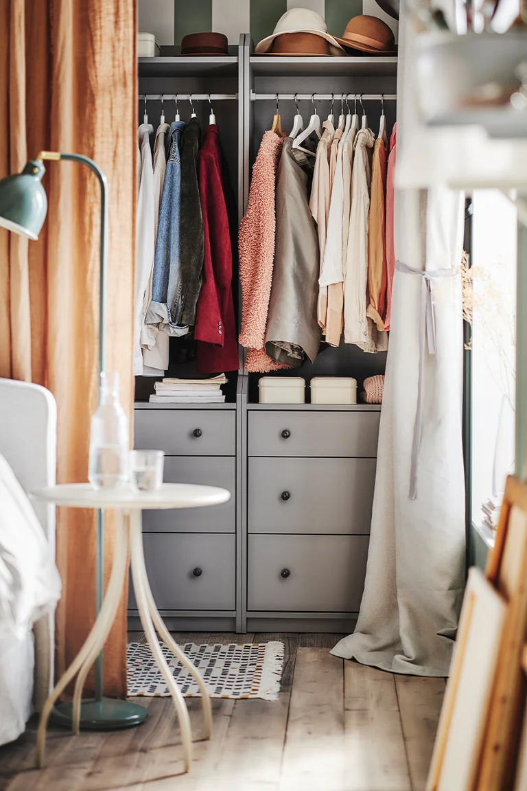 Stunning Kallax Bedroom Transformations You Need to See 4 A bedroom corner with a grey IKEA Kallax wardrobe wall, clothes hanging on rails above drawer inserts, hats on top, a bed edge with white bedding at the left, a small side table with a water carafe, a floor lamp, a patterned rug, and wood flooring