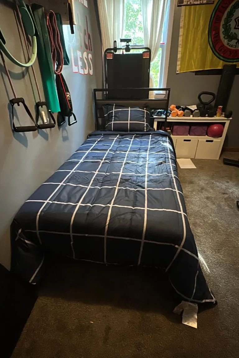 A small bedroom with a single bed in dark blue grid bedding, resistance bands and handles hanging on the left wall, and dumbbells, a kettlebell, and a red ball on a white storage unit beside the bed