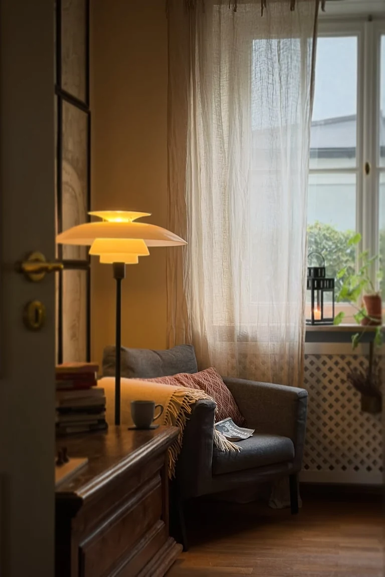 Warm floor lamp glowing beside a gray armchair with a throw in a corner by a curtained window