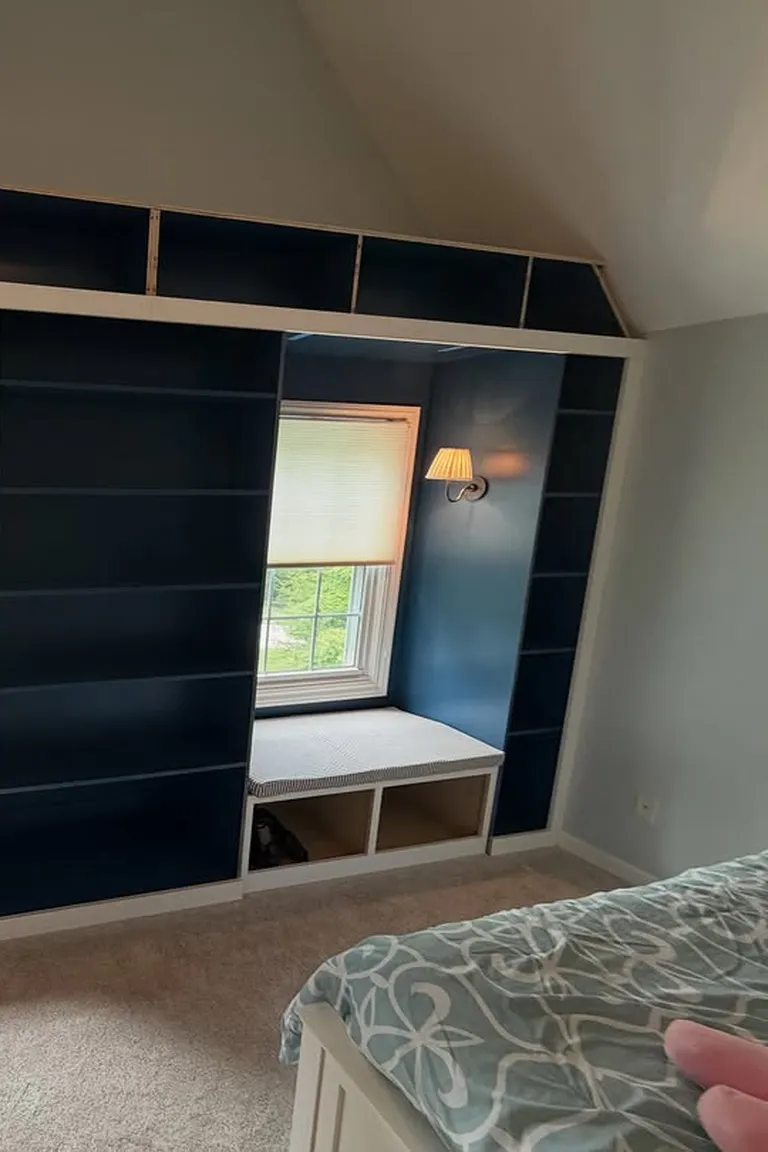 Stunning Kallax Bedroom Transformations You Need to See 5 An attic bedroom with a cushioned window bench and open storage cubbies below, beside a tall dark built-in shelving wall, with the edge of a bed visible in the foreground