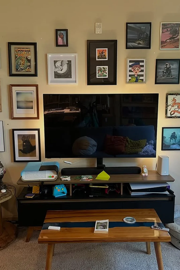 A living room gallery wall with many framed artworks and photos arranged around a large television above a low media console, with a coffee table and rug in front.