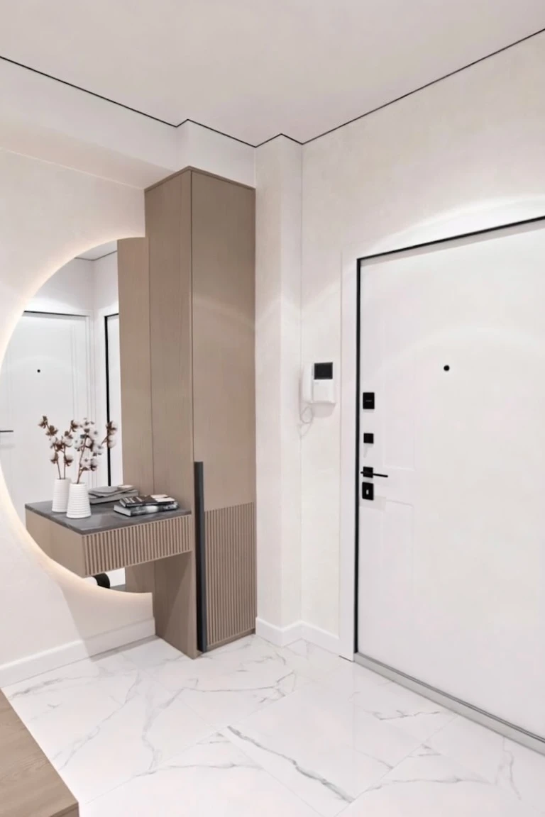 A minimalist white entryway with a tall light wood closed cabinet and a floating console beside a large round backlit mirror, with the white front door visible at right and marble tile flooring