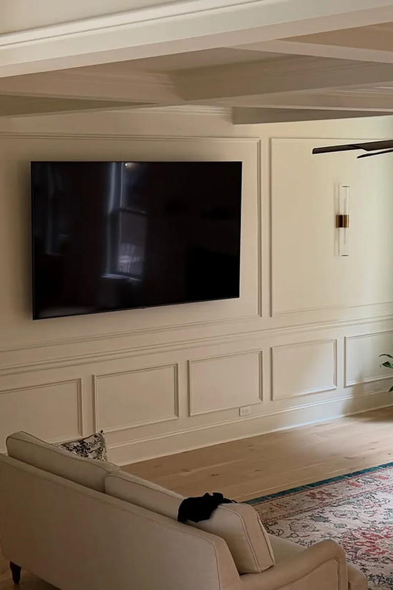 25 Stunning Accent Wall Ideas for Every Room and Style 6 A formal living room with a picture-frame molding accent wall holding a wall-mounted television in the center rectangle, a cream sofa seen from behind, a patterned rug, wood flooring, a window, and a ceiling fan