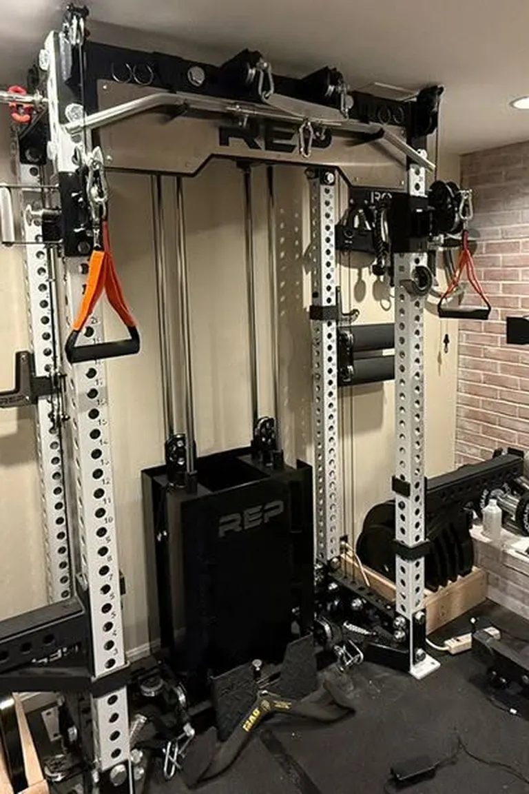 A basement gym with a large silver functional trainer power rack, plate storage, dumbbells, and a brick accent wall on black rubber flooring