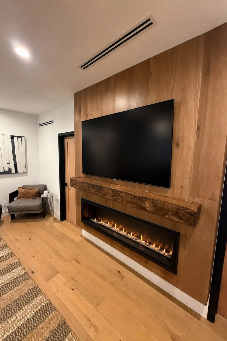 A living room fireplace and TV wall with a large television mounted above a rustic wood mantel and a long linear gas fireplace, set in a wood panelled wall with an armchair and rug nearby.
