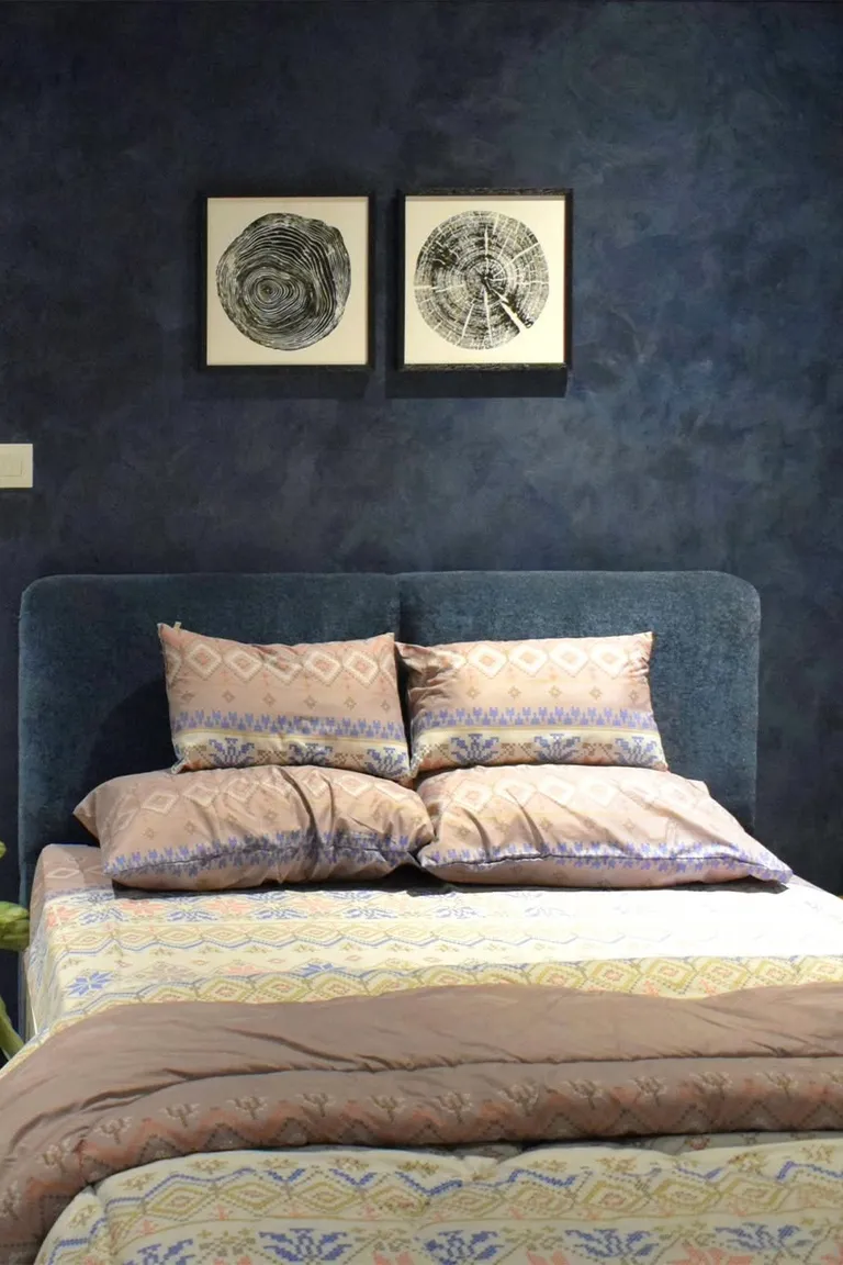 25 Stunning Accent Wall Ideas for Every Room and Style 7 A bedroom with a dark blue limewash plaster accent wall with soft cloudy texture behind a bed with patterned bedding, two framed prints above the headboard, and a plant at the side