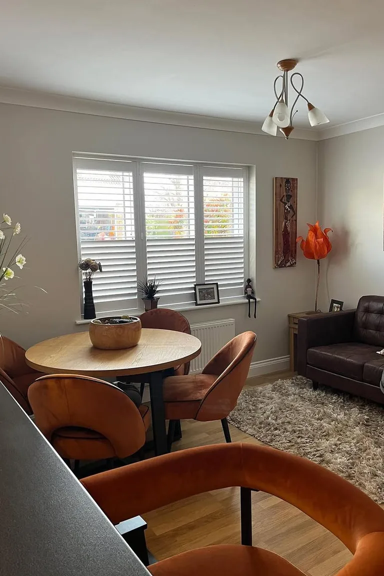 Dining room with a round oak table in front of a full three-panel white plantation shutter window, louvers partly open