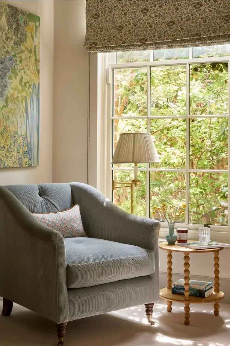 Small two tier side table with books and a candle beside an armchair in a sunny window corner