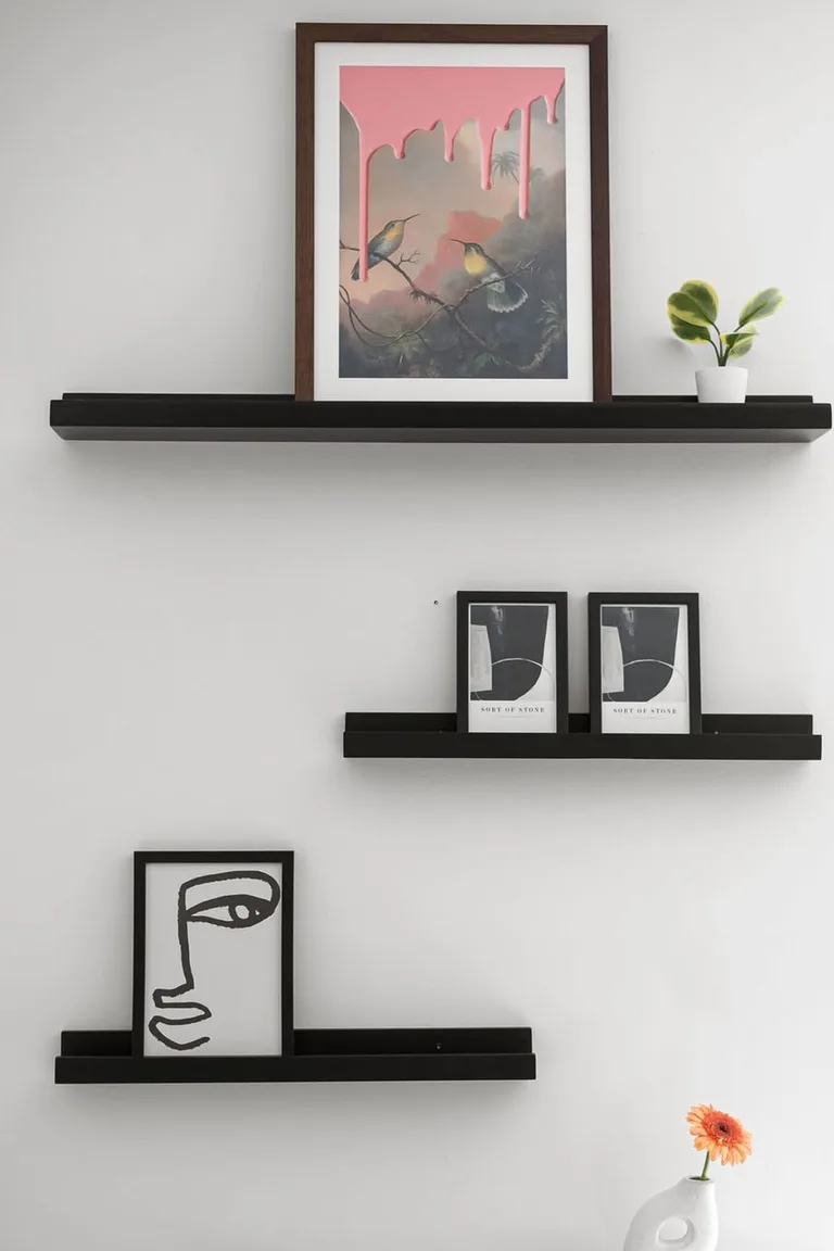 Three staggered black picture ledge shelves on a white wall holding leaning framed artworks and a small potted plant, with a white vase and single flower cropped at the bottom edge