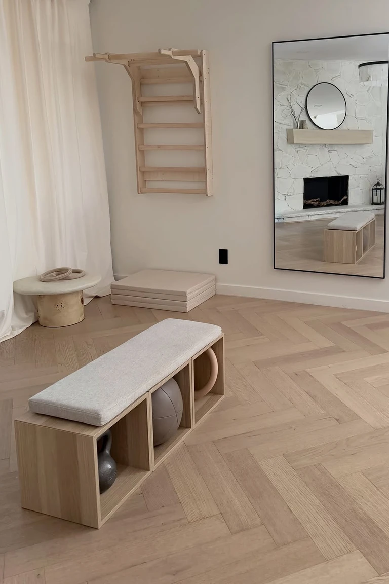 A bright minimalist workout room with herringbone wood flooring, a tall leaning mirror, a wooden wall-bar unit, a low storage bench, and folded mats