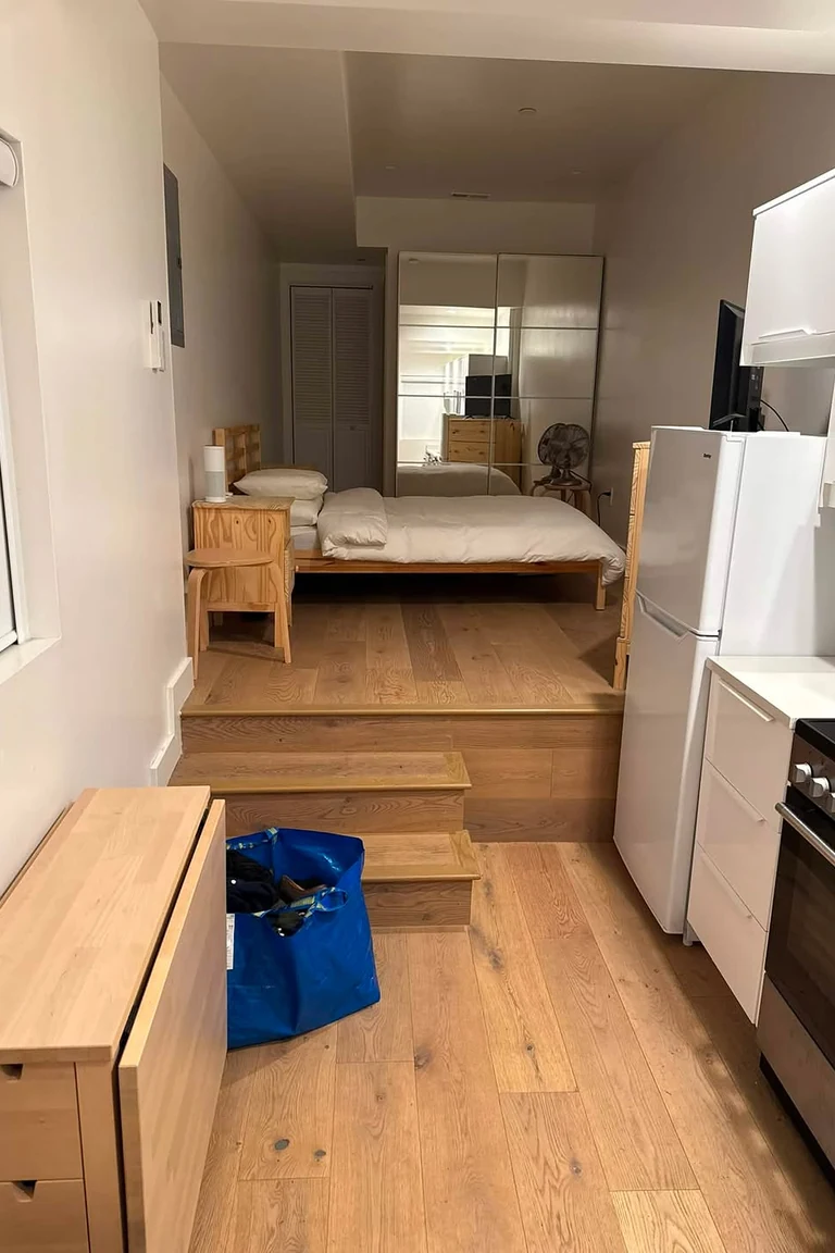 11 Micro Studio Apartment Ideas for Extremely Small Urban Spaces 8 Micro studio with a raised wood platform bed with steps, white kitchenette with fridge and oven at the right, and wood floors