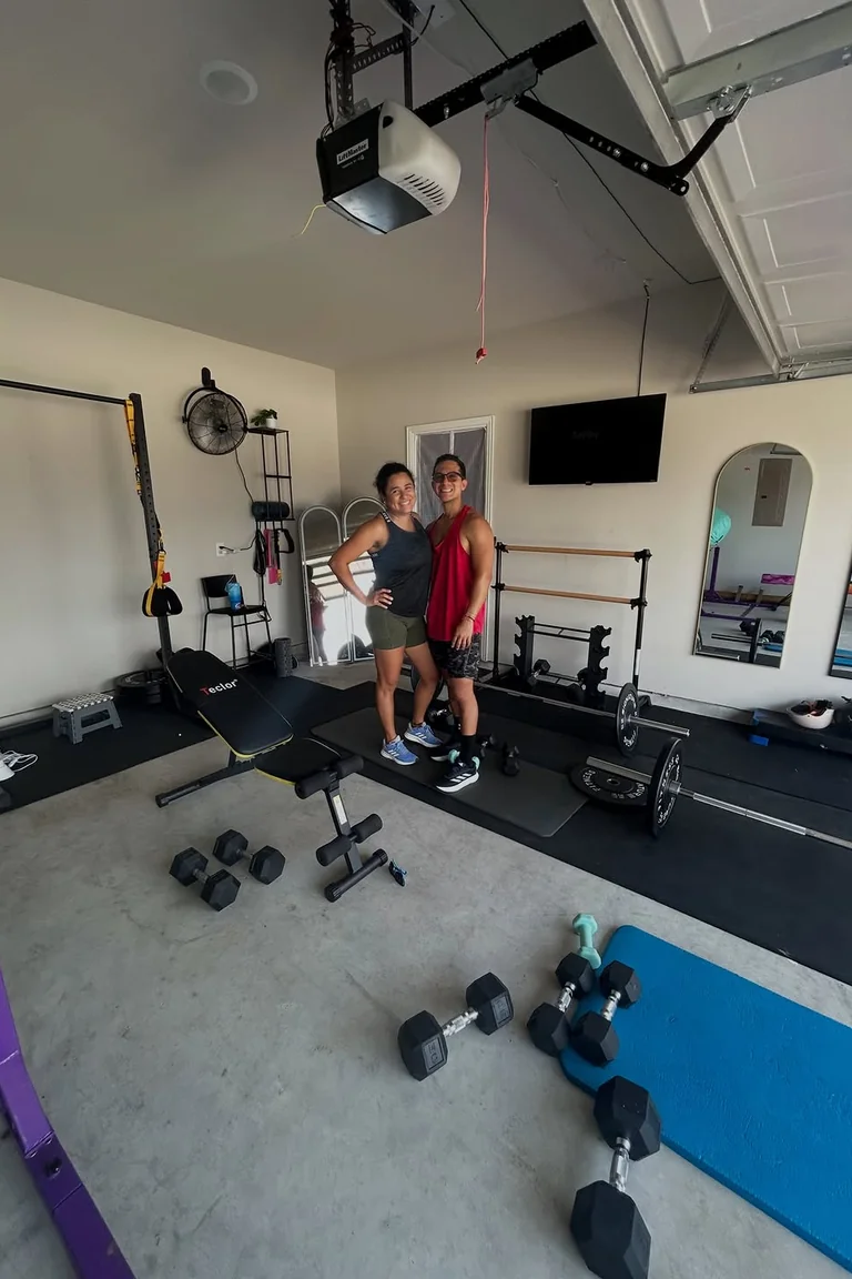 A bright converted garage gym with white walls, black rubber mats, a weight bench, dumbbells, barbells, a wall-mounted TV, arched mirrors, and a garage door opener overhead, with two people posing in the center