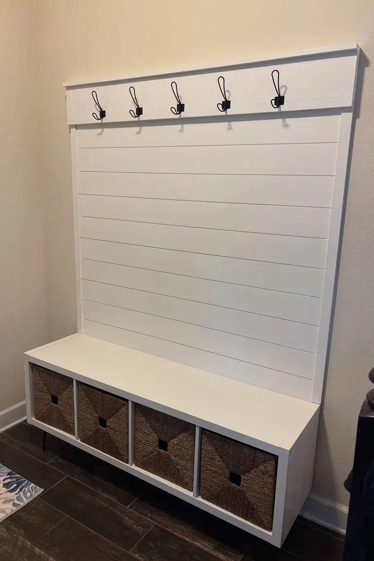 A white entryway bench unit with a shiplap back panel, five black coat hooks above, a bench seat, and four woven baskets in cubbies below, on a dark wood floor