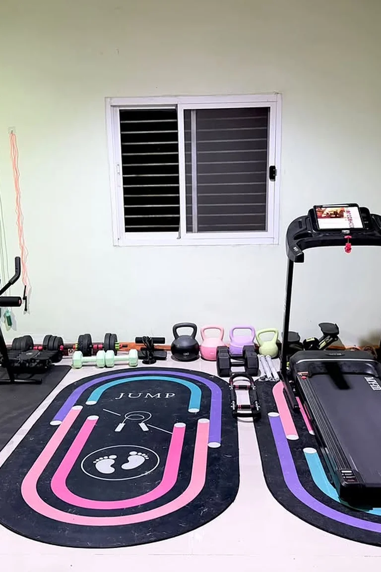 A home fitness room with pale green walls, a treadmill on a colorful pink and blue mat, colorful kettlebells and dumbbells lined up along the wall, and an ab trainer on a black mat