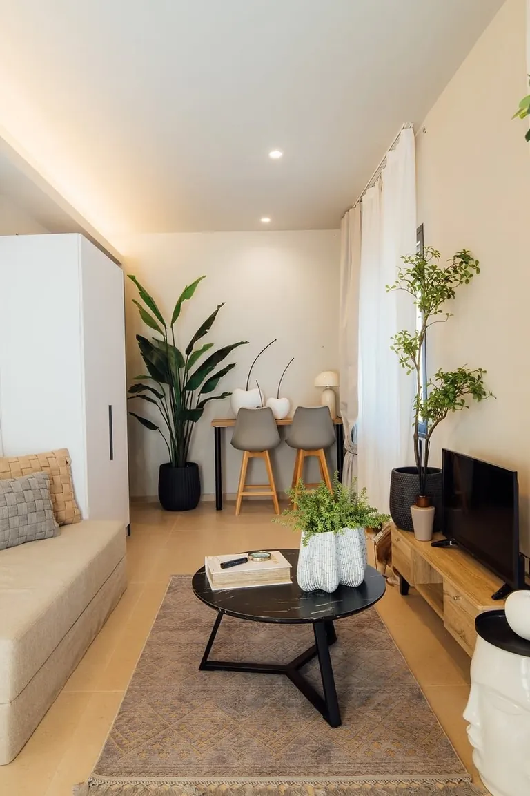 11 Micro Studio Apartment Ideas for Extremely Small Urban Spaces 10 Small studio living room with a beige daybed with cushions at the left, a round coffee table, a desk with chairs, and a TV console