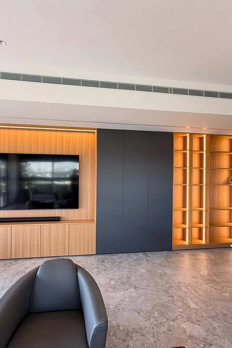 An open-plan living and dining area with a TV wall of wood panelling, low cabinets and lit shelving, armchairs in front and a dining table with pendant light to the side.
