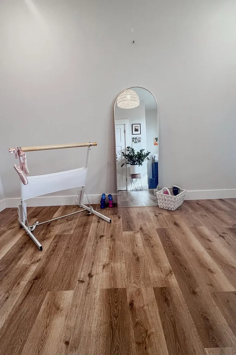 A home barre corner with a white portable ballet barre holding pink ballet shoes, a tall arched leaning mirror, small dumbbells, and a woven basket of gear on a wood floor