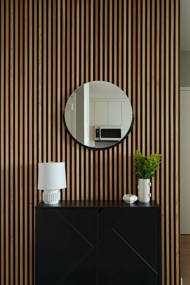 A floor-to-ceiling vertical wood slat accent wall with a round black-framed mirror above a black console cabinet holding a white lamp, small bowl, and vase of greenery