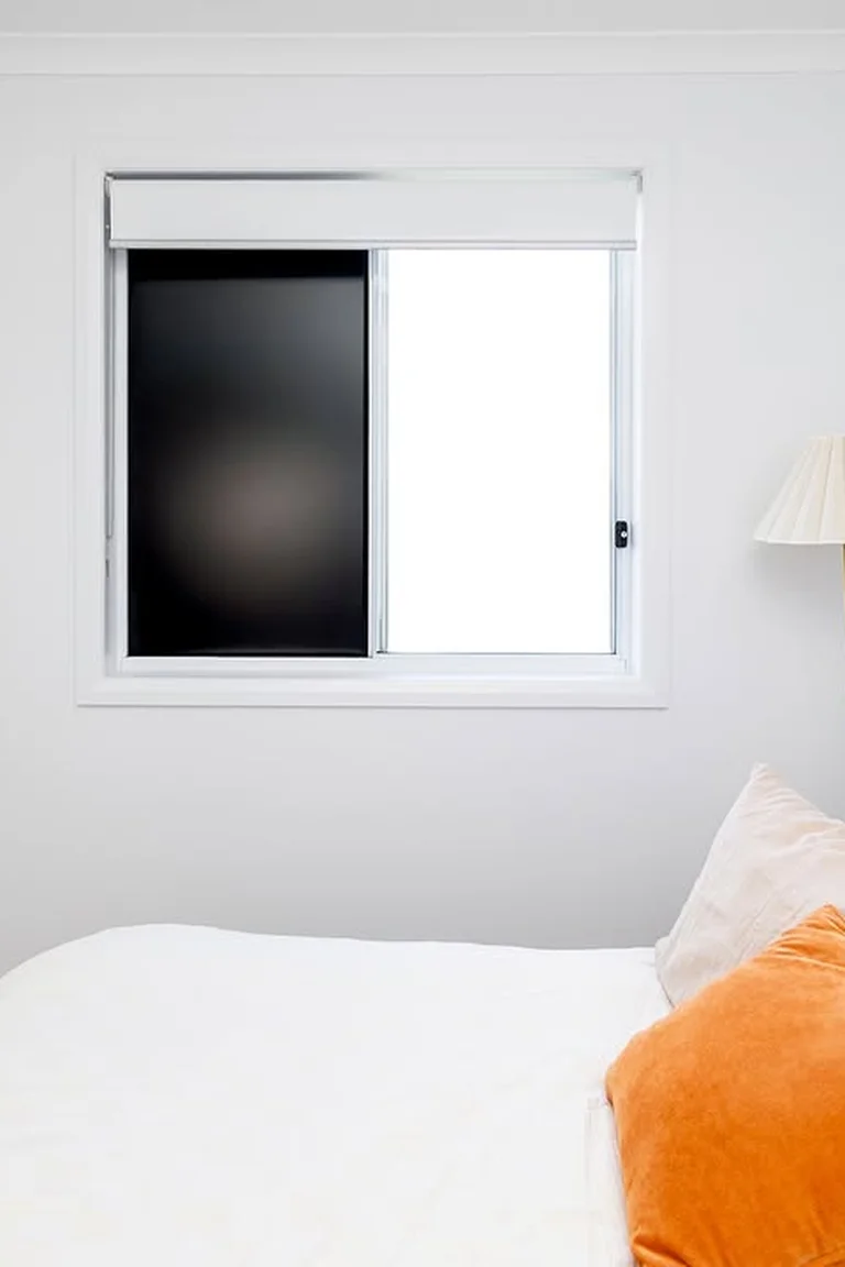 Tidy bedroom with a sliding window, one pane covered by a solid black blackout panel, made bed with burnt-orange cushion