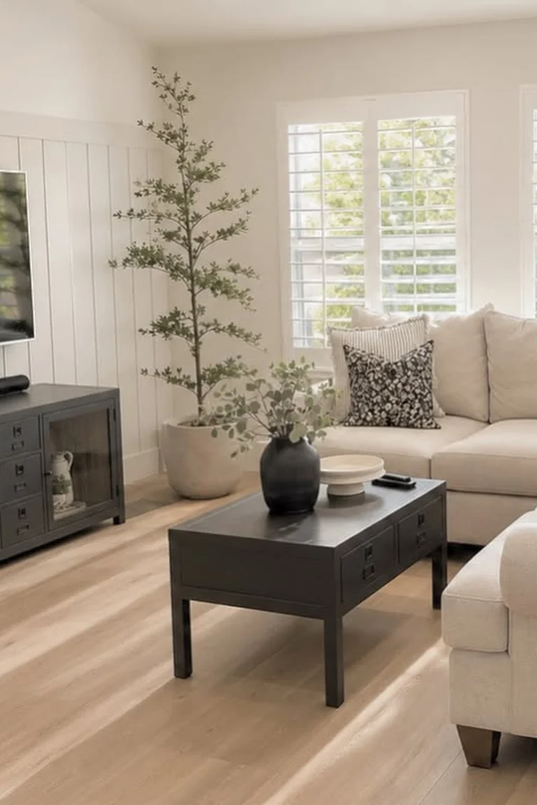 A bright farmhouse living room with a white vertical shiplap TV wall, a large television above a dark console, a beige sectional sofa, a coffee table and shuttered windows.