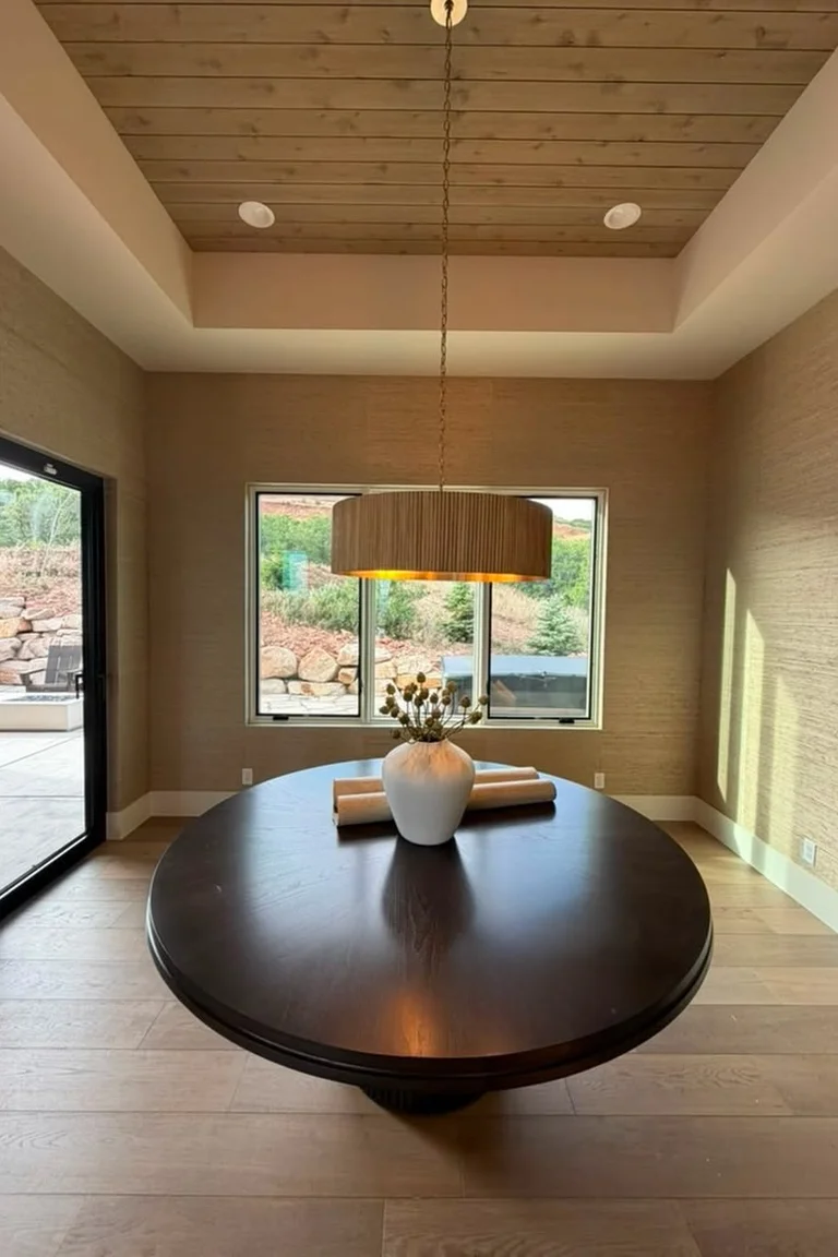 25 Stunning Accent Wall Ideas for Every Room and Style 13 A dining room with natural beige grasscloth wallpaper on the walls around a large round dark wooden table with a white vase, a drum pendant light, wood flooring, and windows looking onto a garden