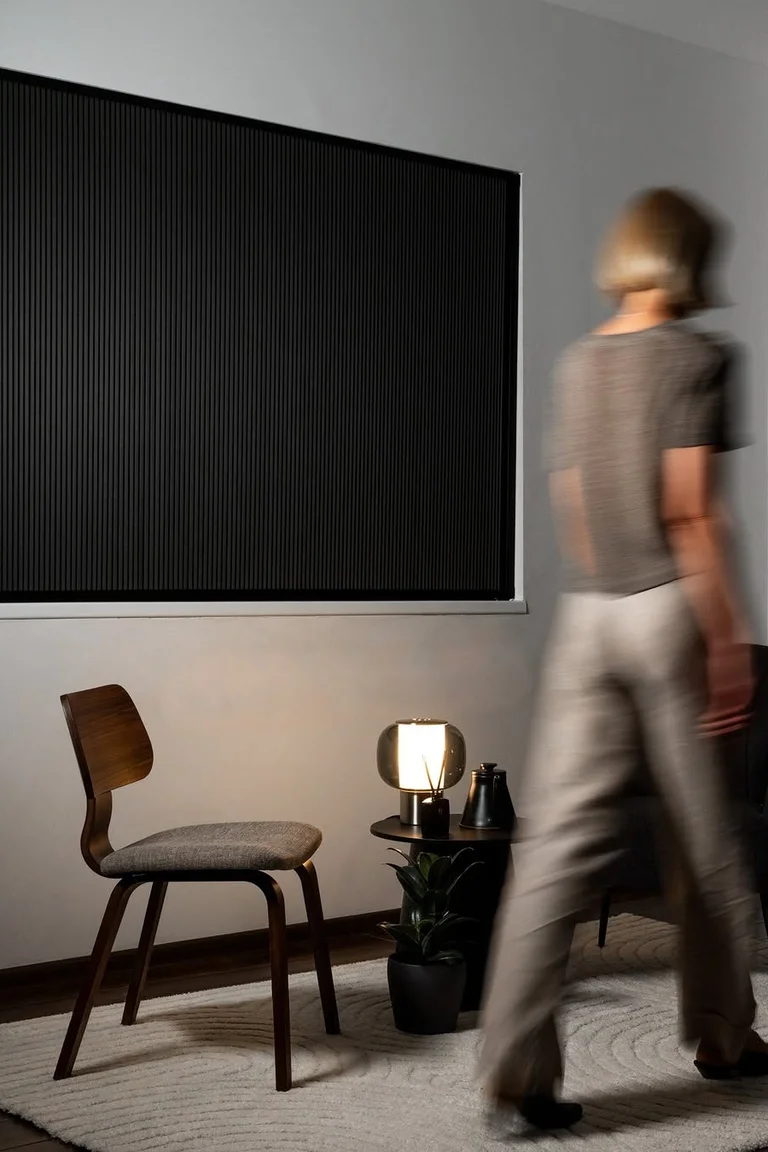 Dim room with a large window fully covered edge-to-edge by a black pleated panel, chair and lit lamp below