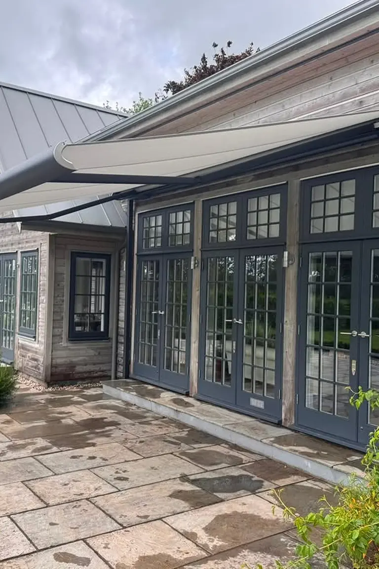 Timber house with a large beige retractable awning fully extended over French doors and windows, stone patio and garden