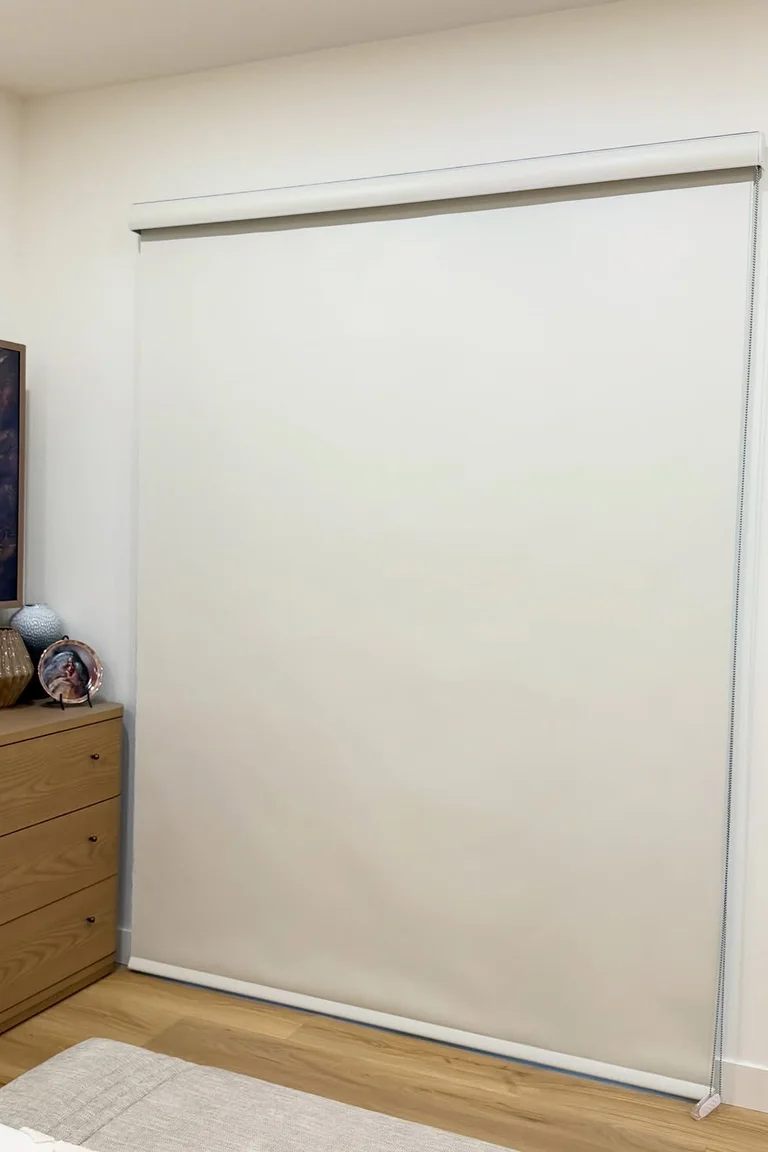 Bedroom corner with a white blackout roller shade fully lowered over a sliding glass door, wood dresser and bed corner