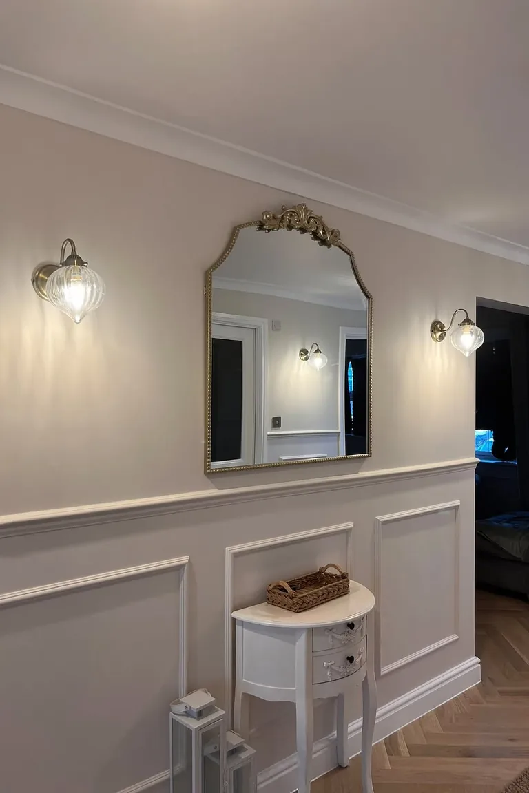Hallway wall with white panel molding, an ornate gold mirror, brass sconces, and a small white console
