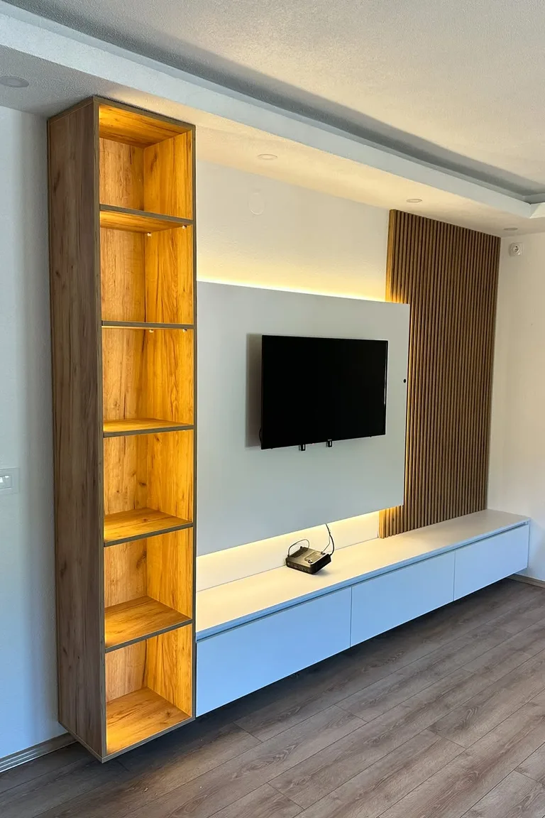 A narrow living room TV wall with a tall illuminated wooden shelving unit beside a wall mounted television and a long low white console.