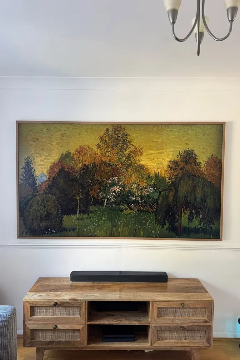 A Samsung Frame style television displaying a landscape painting in a slim wood frame above a wooden console in a living room.