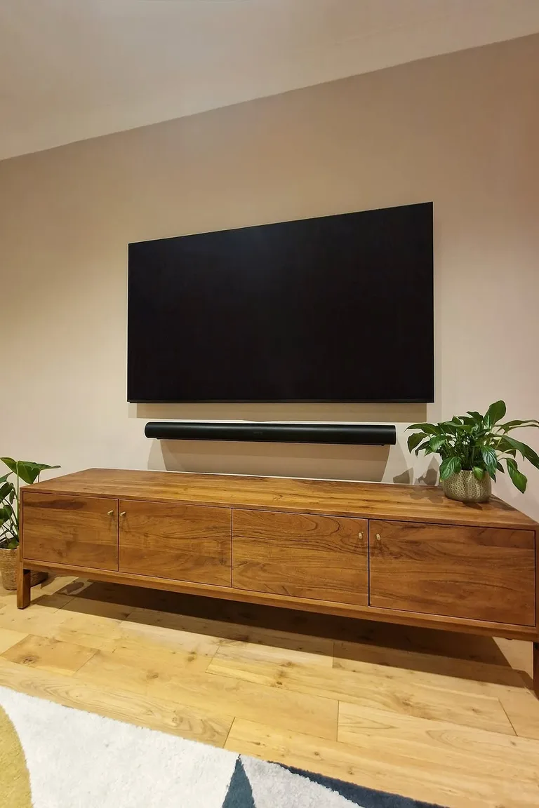 A wall mounted television with a soundbar above a simple long wooden console on legs against a warm painted wall.