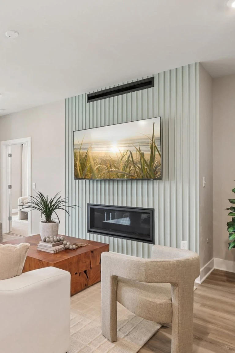 Bright living room with a light sage vertical slat accent wall, a wall-mounted TV above a linear fireplace, beige armchairs, a wooden coffee table, plants and wood flooring
