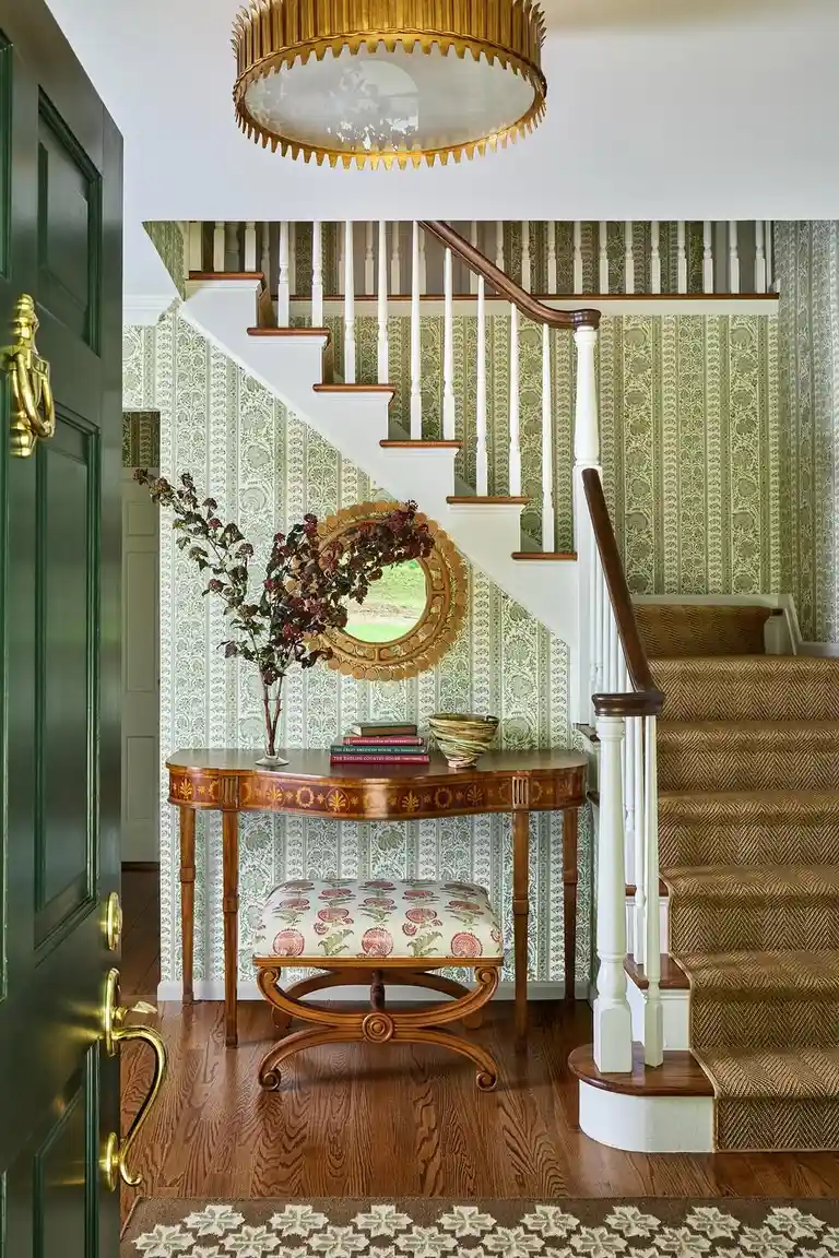 A welcoming entryway viewed from just inside an open dark green front door with brass hardware, showing a wood console table with books and bowl, round gold mirror, patterned green wallpaper, staircase with wood handrail, and hardwood floor with patterned rug