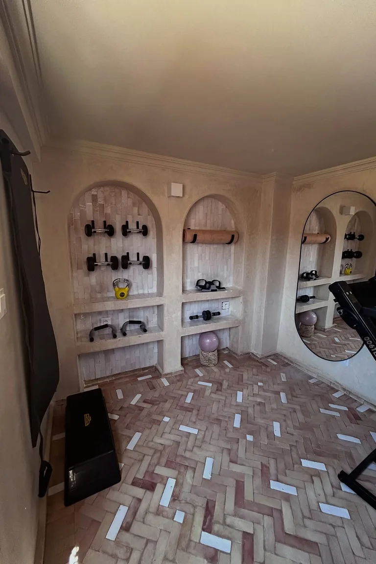 A small stylish home gym room with arched wall niches holding dumbbells and a kettlebell, a large oval mirror, a step platform, and herringbone tile flooring