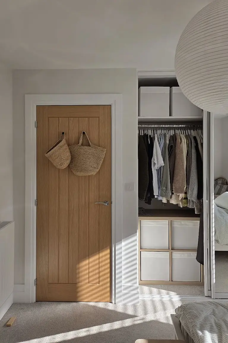Bright bedroom with a wooden door, an open wardrobe with white Kallax cube boxes below hanging clothes, a paper pendant lamp, and the edge of a striped bed in the foreground